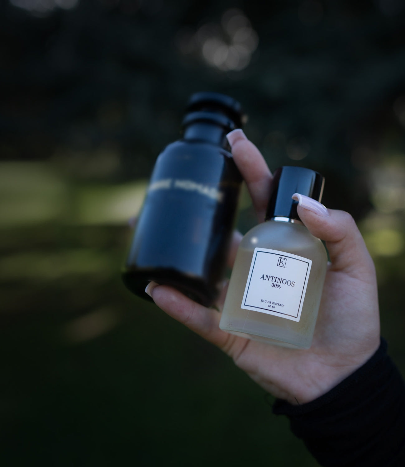 Eine Hand hält zwei Flaschen: eine schwarze, undurchsichtige Flasche mit unleserlichem Etikett sowie eine klare Flasche mit schwarzem Verschluss und weißem Etikett, auf dem „ANTINOOS 30% EAU DE EXTRAIT“ steht. Im Hintergrund ist unscharfes natürliches Grün zu sehen.
