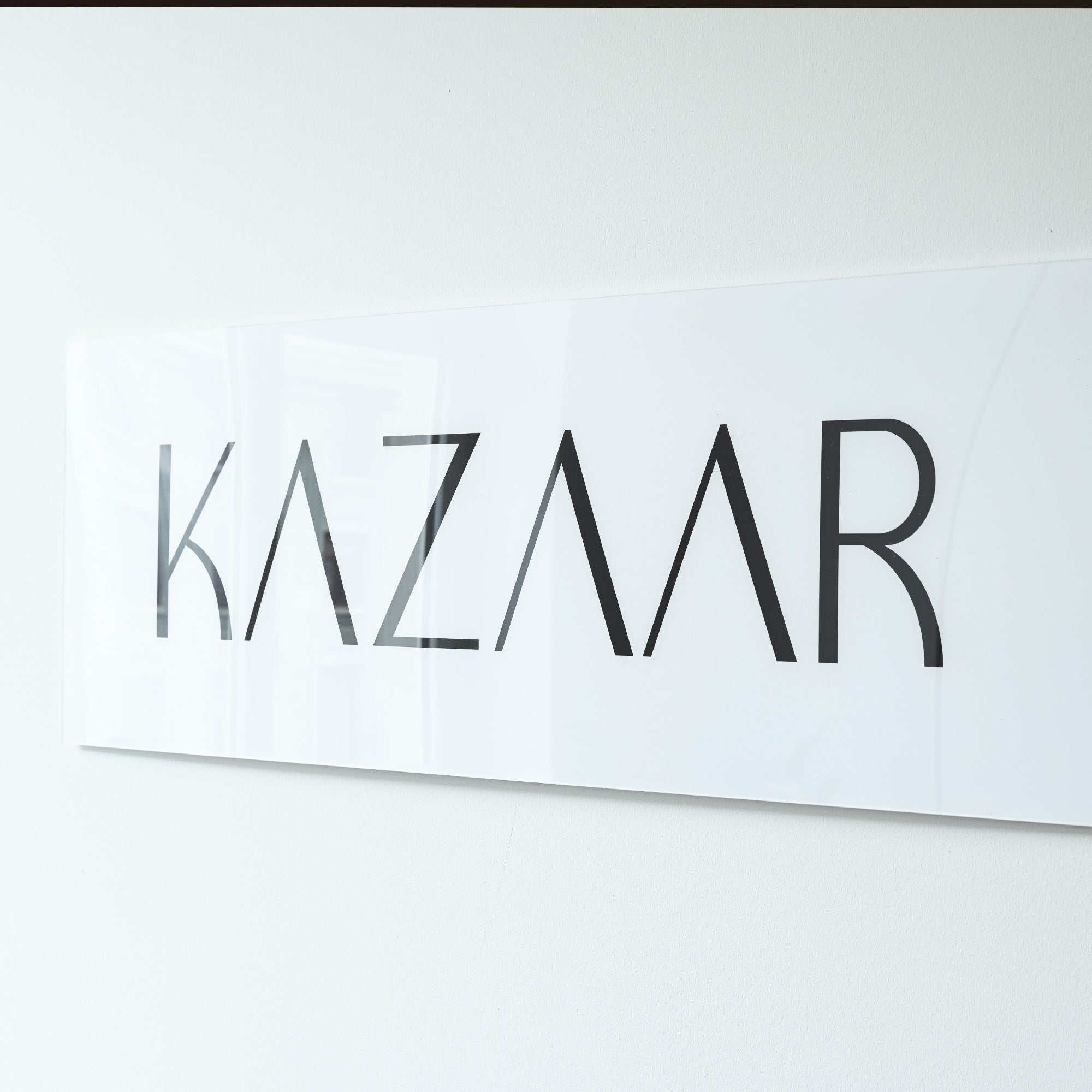 Ein rechteckiges, glänzendes Schild an einer weißen Wand mit schwarzer, moderner Schrift "KAZAAR" in Großbuchstaben.