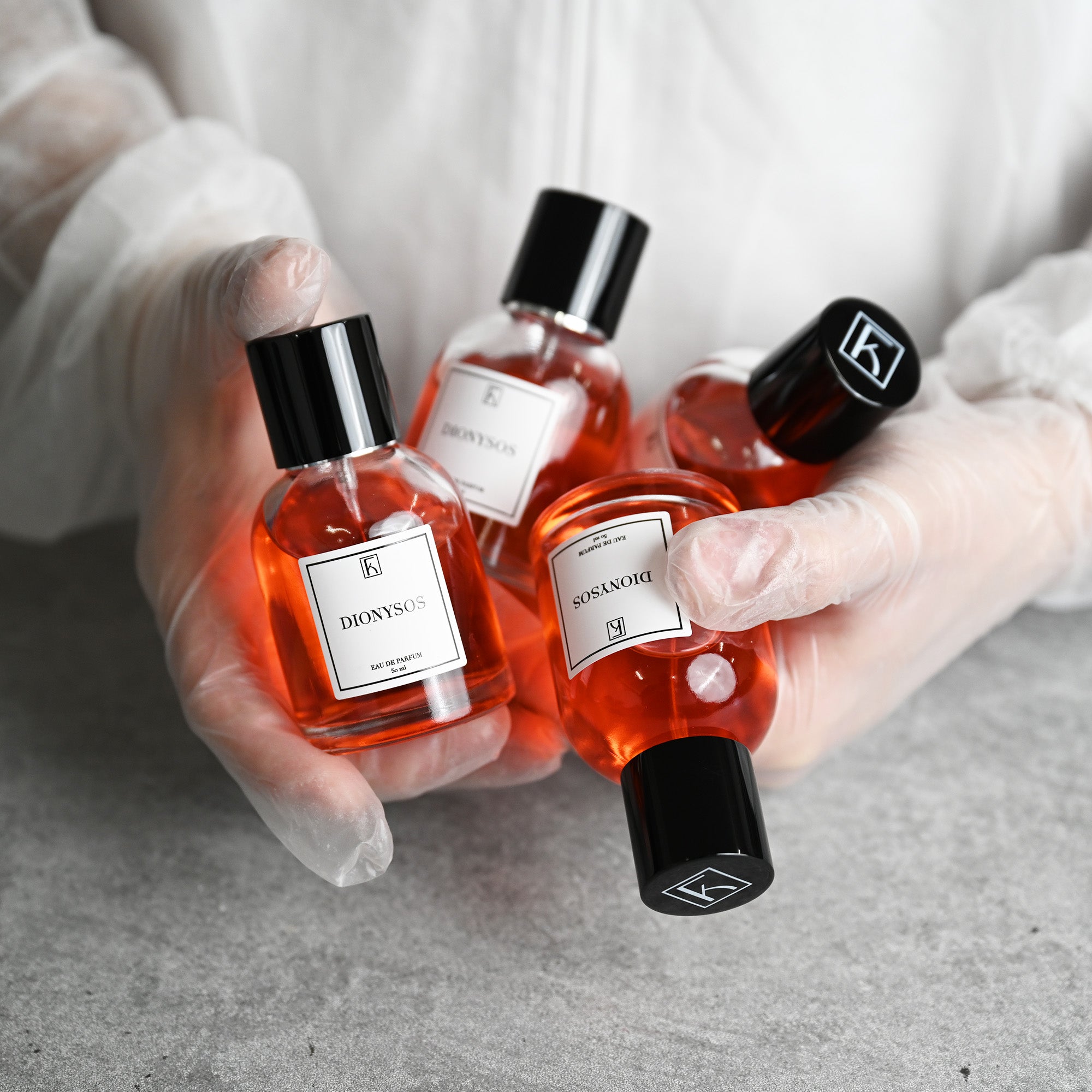 Eine Person mit weißen Handschuhen hält mehrere Flaschen „DIONYSOS“ Eau de Parfum mit orangener Flüssigkeit und schwarzem Deckel.