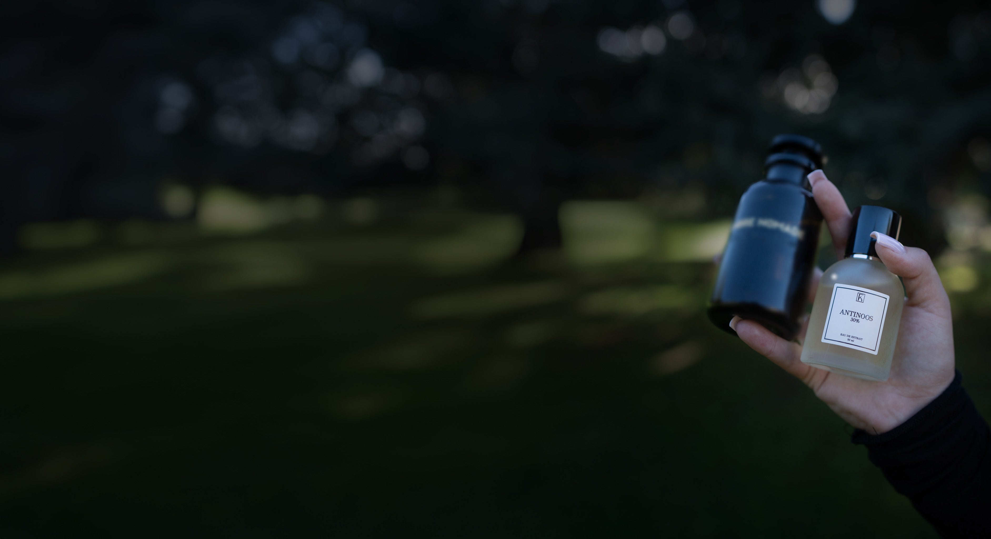 Eine Hand hält zwei kleine Flaschen, eine schwarz und undurchsichtig, die andere durchsichtig mit hellem Inhalt und dem Etikett "ANTINOOS 30%". Im Hintergrund ist eine grüne, unscharfe Wiese mit Bäumen zu sehen, Tageslicht.