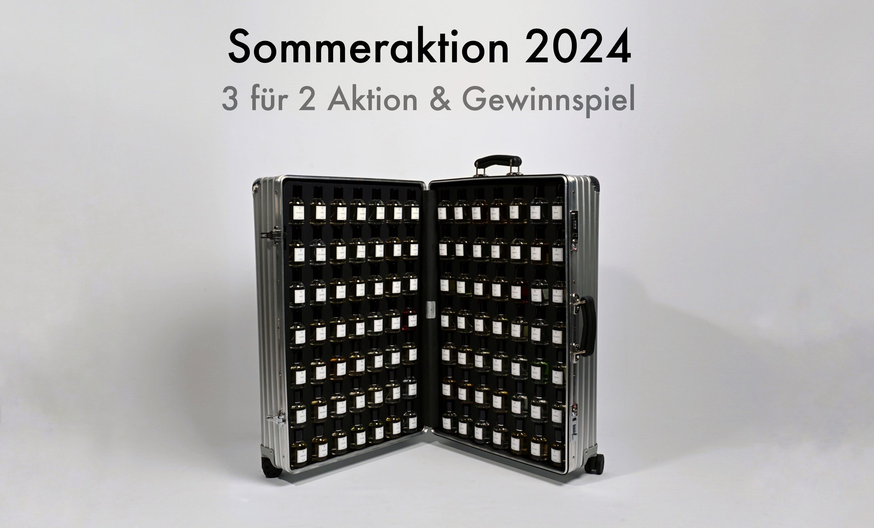Auf dem Bild ist ein silberner Koffer zu sehen, der offen steht. Im Inneren befinden sich viele kleine, braune Fläschchen, jeweils in einzelnen Fächern einsortiert. Alle Fläschchen haben weiße Etiketten. Der Koffer steht auf einem hellen Untergrund. Oben im Bild steht groß „Sommeraktion 2024“, darunter kleiner „3 für 2 Aktion & Gewinnspiel“. Sonst sind keine weiteren Objekte oder Personen erkennbar.