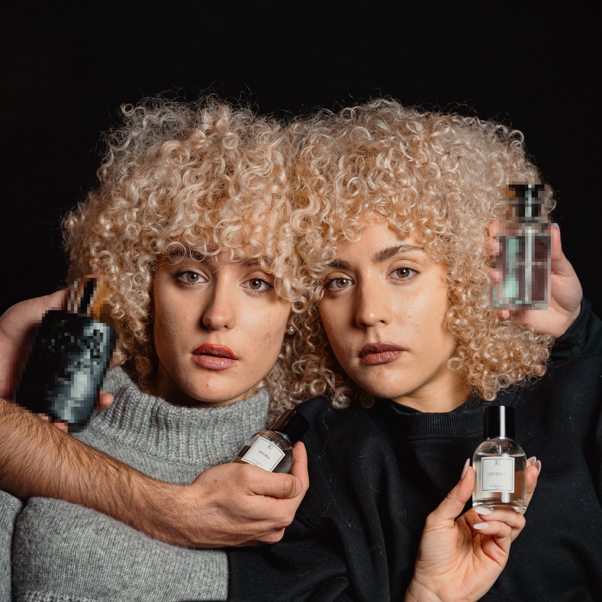 Zwei Frauen mit lockigem, blondem Haar stehen nebeneinander vor schwarzem Hintergrund. Beide halten Parfümflaschen in den Händen, eine Person reicht von links und rechts jeweils eine weitere Flasche ins Bild. Die Frauen tragen dunkle Pullover. Die Etiketten auf den Parfümflaschen sind teilweise lesbar. Im Vordergrund stehen die Gesichter der Frauen und die Flaschen im Fokus. Die Szene wirkt wie eine Werbeaufnahme für Parfums.