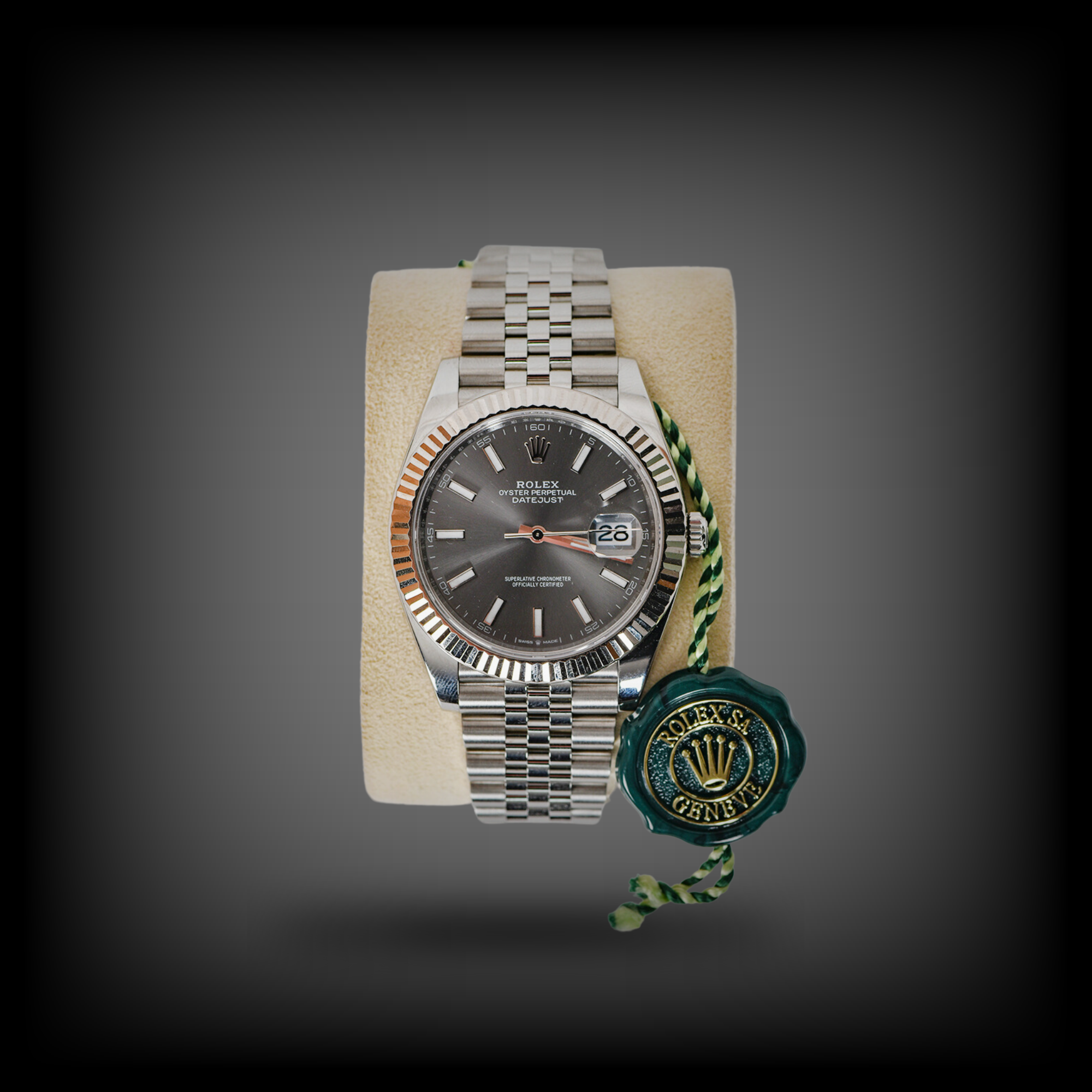 Eine silberne Rolex-Armbanduhr mit dunklem Zifferblatt und Datumsanzeige liegt auf einem beigen Kissen, daneben ein grünes Siegel.