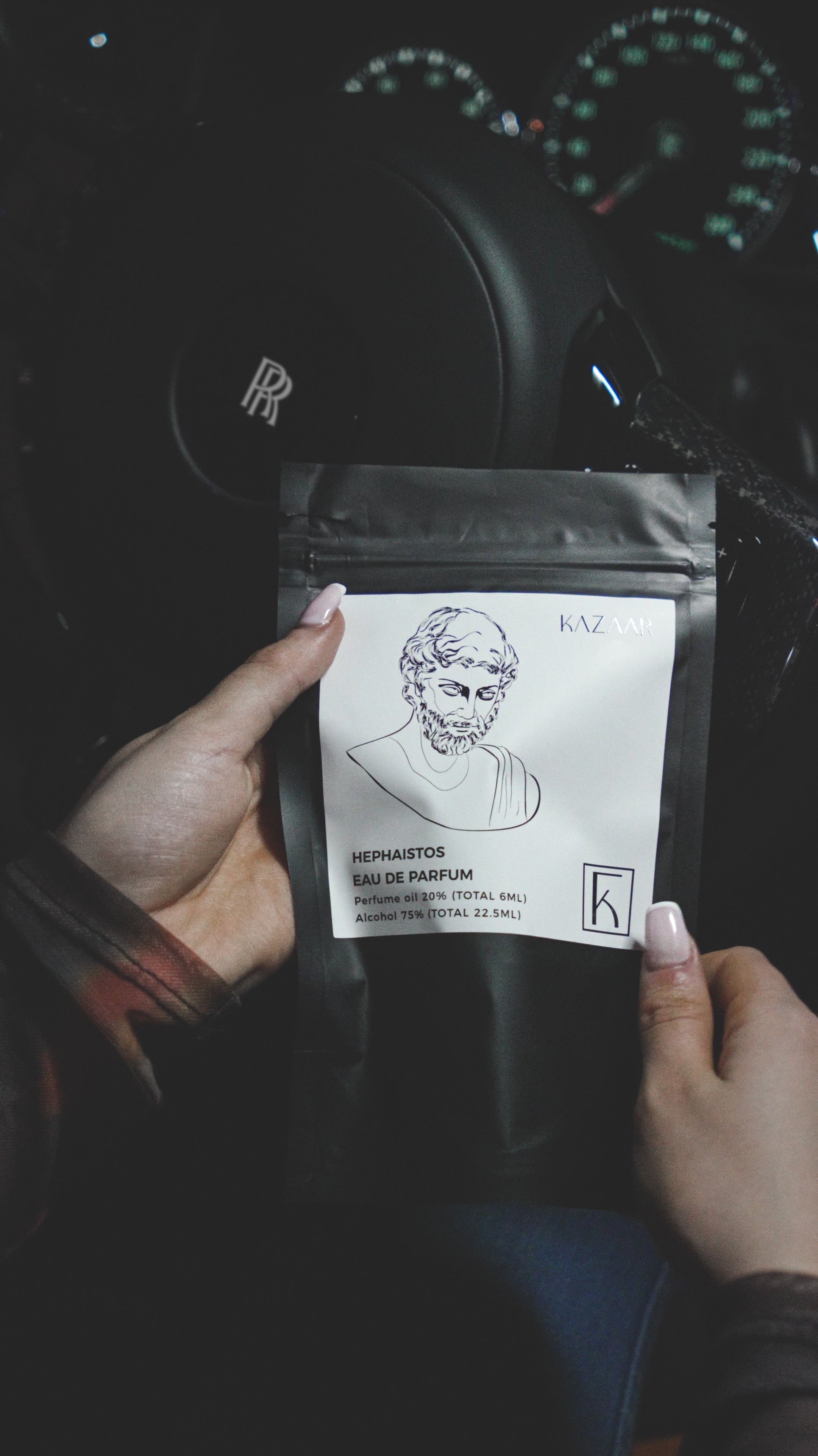 Eine Person mit lackierten Fingernägeln hält eine schwarze Verpackung mit weißem Etikett, auf dem ein gezeichnetes Porträt eines bärtigen Mannes und die Aufschrift „HEPHAISTOS EAU DE PARFUM“ zu sehen sind. Das Etikett enthält weitere Informationen zur Parfümöl- und Alkoholkonzentration. Die Verpackung trägt das Logo „KAZAAR“ und ein stilisiertes „K“. Im Hintergrund sieht man das Lenkrad eines Autos mit Rolls-Royce-Emblem sowie verschwommene Armaturen auf dem Armaturenbrett.