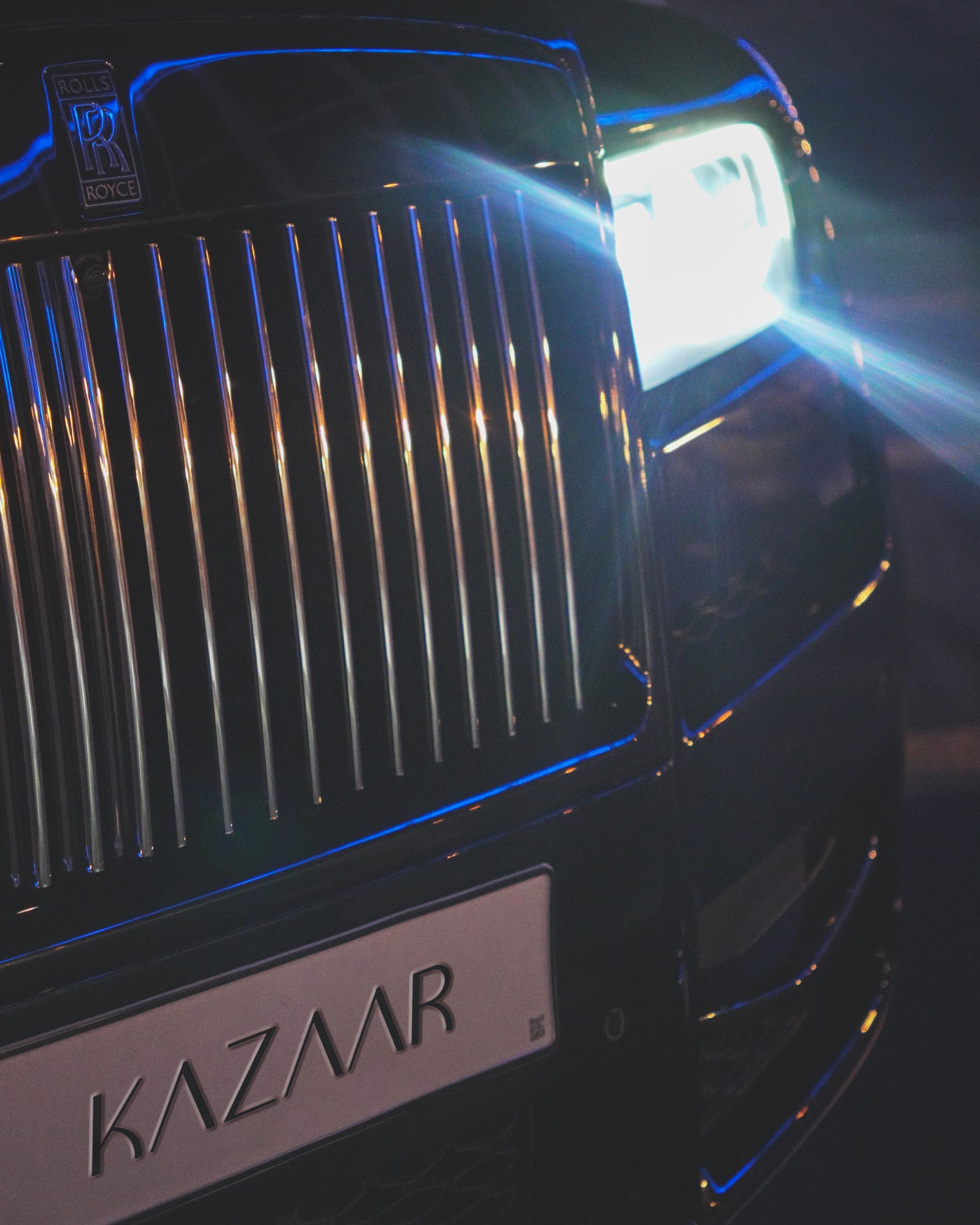 Im Bild ist der vordere Bereich eines Luxusautos im Dunkeln zu sehen, darunter ein markanter Rolls-Royce-Kühlergrill mit vertikalen Chromstreben. Am oberen linken Rand befindet sich das Rolls-Royce-Emblem. Rechts im Bild leuchtet ein heller LED-Scheinwerfer, dessen Licht einen deutlichen blauen Lichtstrahl erzeugt. Unten am Auto ist ein Kennzeichen mit der Aufschrift „KAZAAR“ zu erkennen. Die Karosserie reflektiert blaue und orangefarbene Lichter aus der Umgebung.